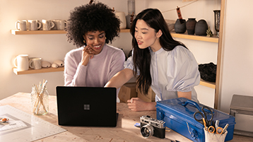 Two women collaborating at a table with a laptop in a pottery studio, surrounded by ceramics, tools, and a camera.