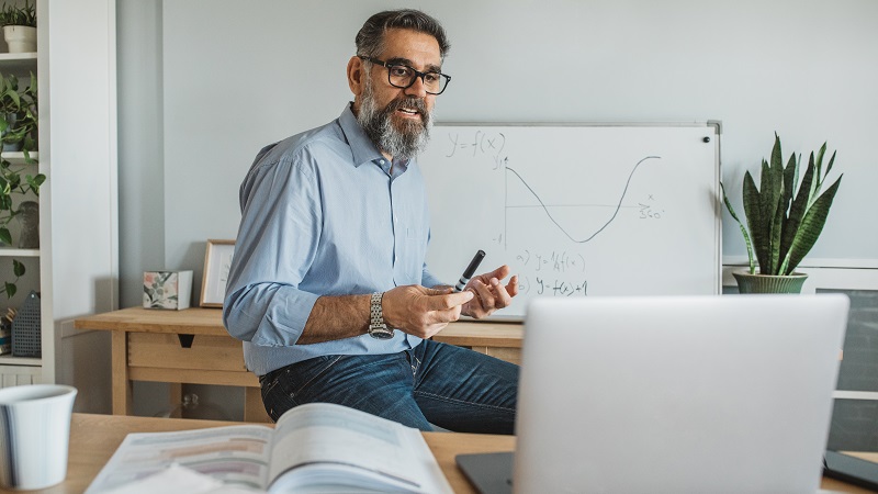 Photo of a teacher at a laptop with a whiteboard behind him.